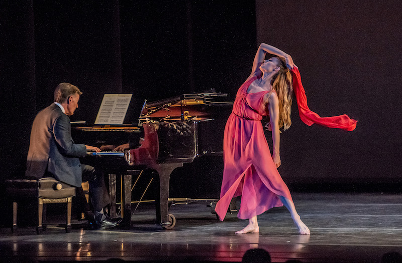 A man in a suit sits at a grand piano. Sara Mearns, a coral grecian dress, stands near him. She waves a pink scarf that cascades near her face.
