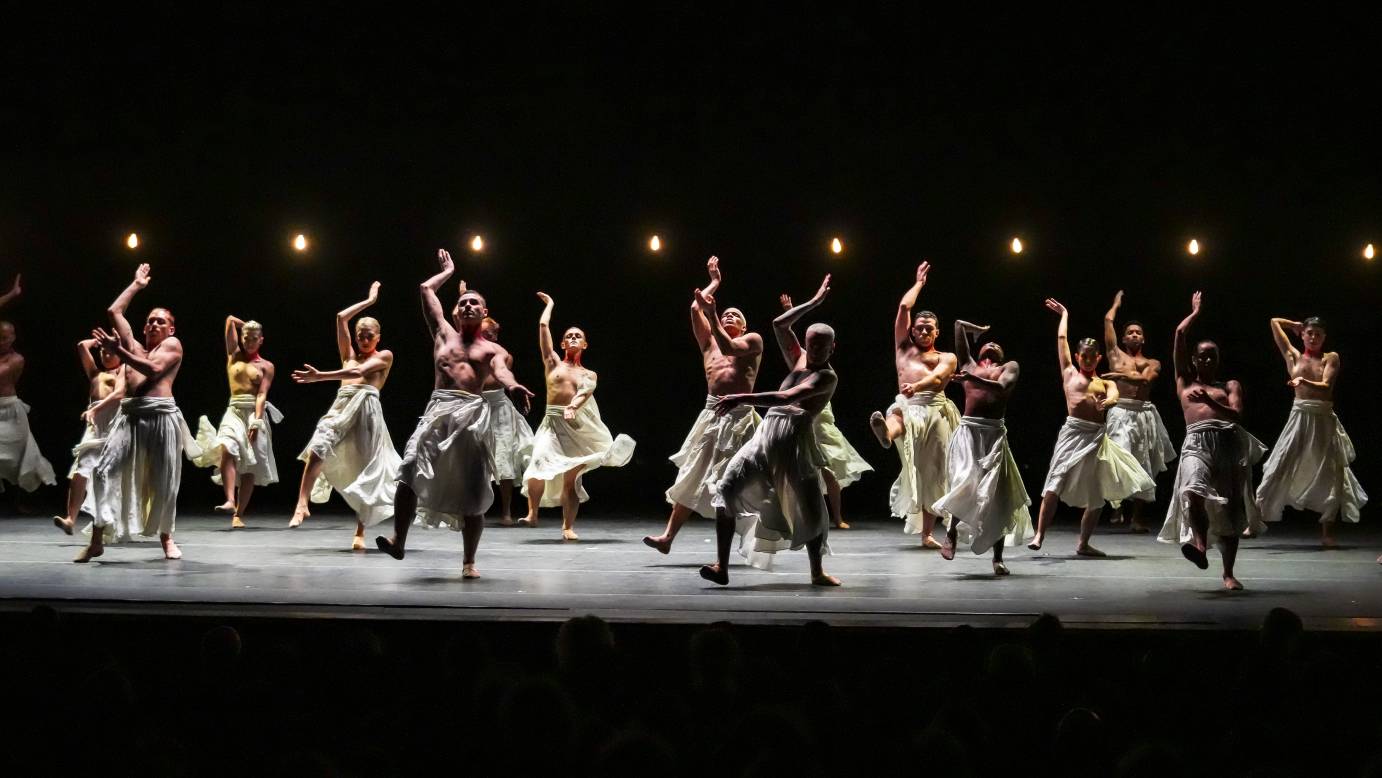 a group of 20 topless dancers, men and women, wearing sheer white skirts balance on one leg and lean back as they wave their arms