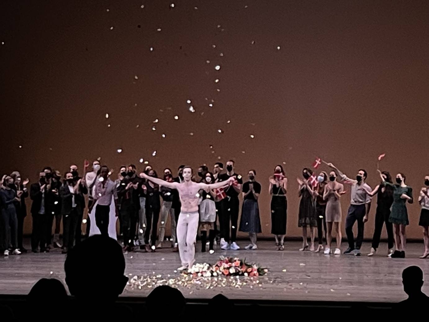 Barechested, Ask la Cour stands in a pile of flowers, his arms raised. Other dancers stand behind him, applauding.