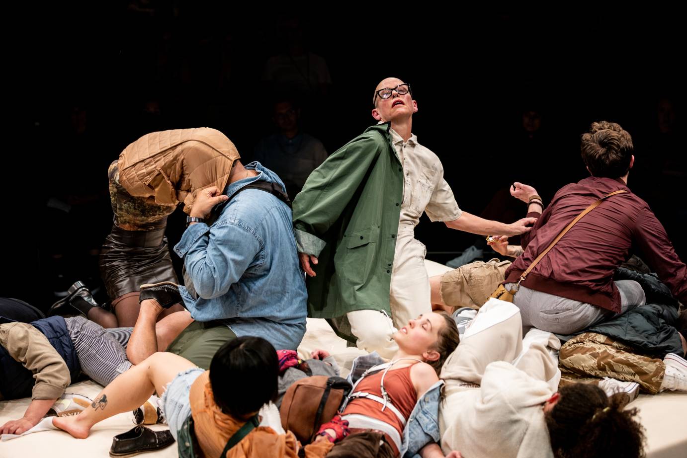 Intertwined performers atop a spongy white platform. A central figure is a white shaved head woman wearing black framed glasses and a green canvas coat, a kneeling man wearing a blue denim shirt with a quilted gold coat over his head and a seated white brown haired man with his back to us in brown coat. At least four other performers in various states of undress and splayed on the platform