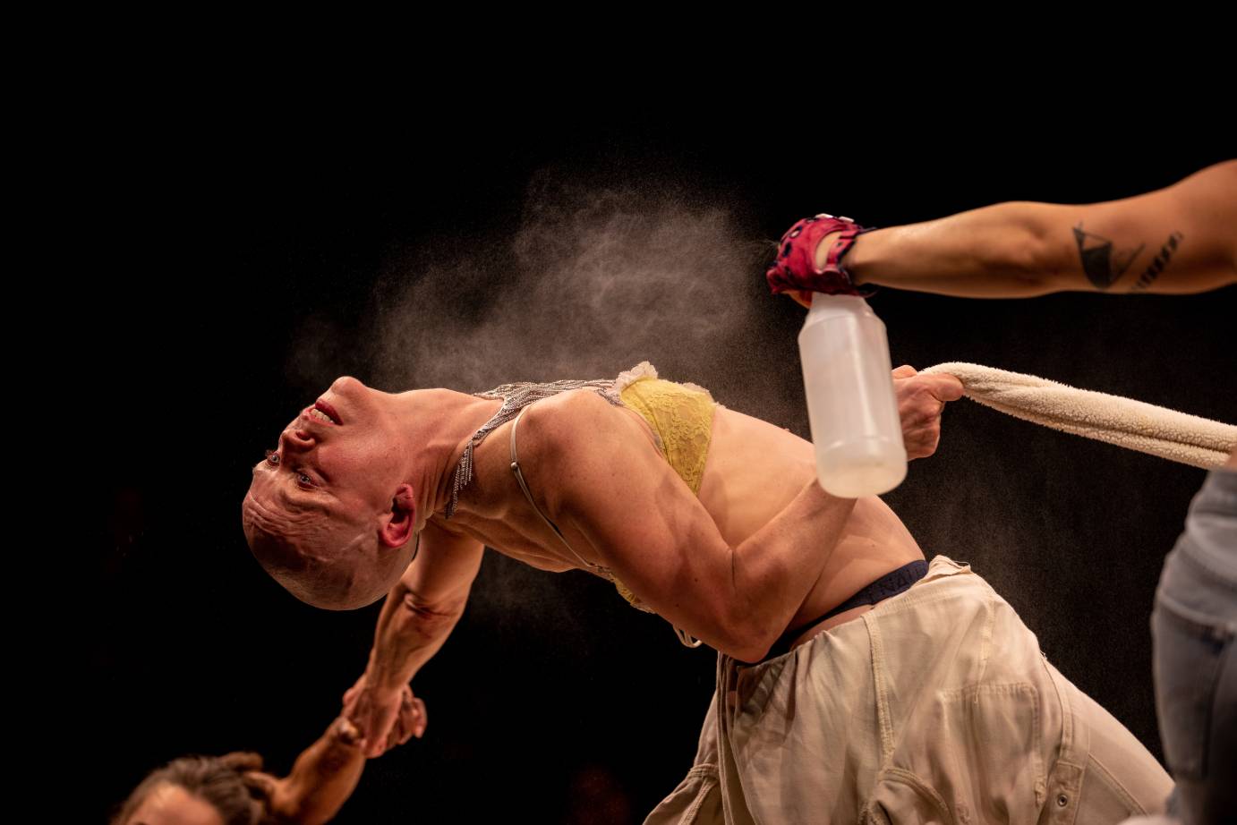 Red gloved hand holds a spritz bottle that sprays an extremely arched white woman seen in profile wearing a greenish bra and white flowing shorts