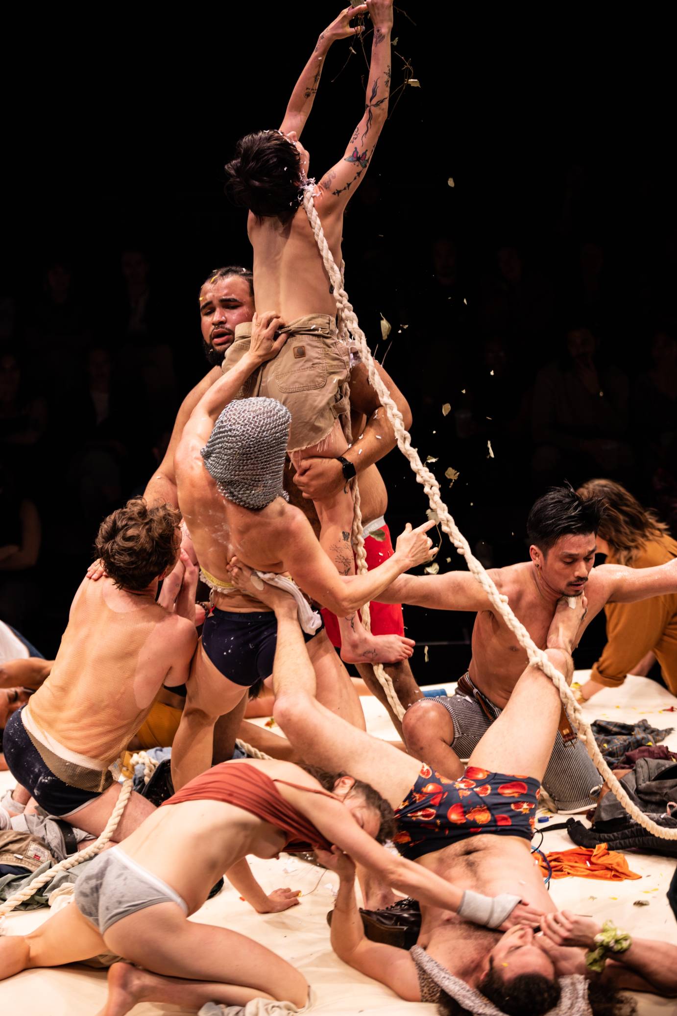 A white woman with arms stretched to the ceiling crumbles leaves as she is lifted by performers balanced on a white spongy platform