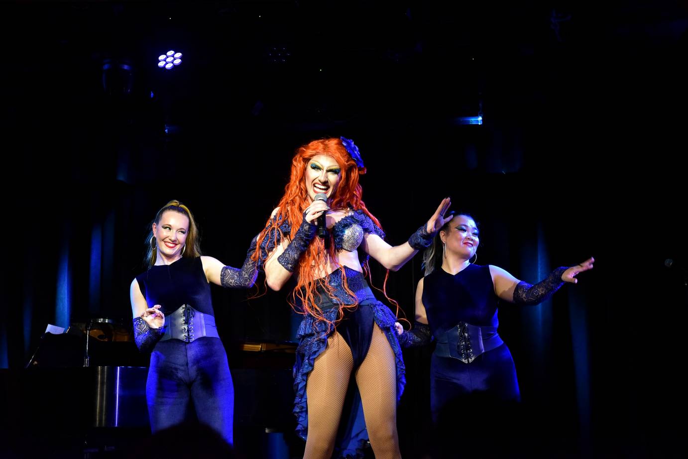 Drag queen in red wig and sparkly blue 'maritime-esque' costume with two women back up singers in blue, all gesturing with left hands on left diagonal, elbows bent