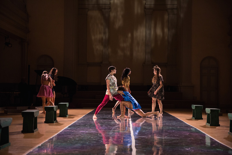 Three dancers turn to walk while another kneels between them. Two other dancers embrace off to the side.