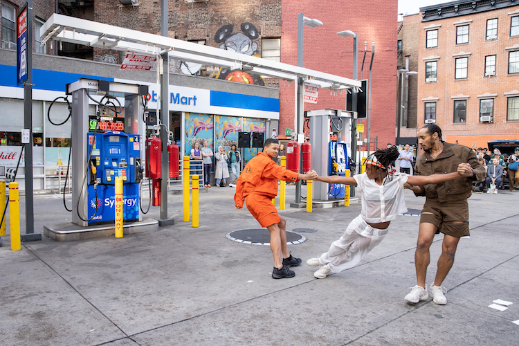 three dancers one in an orange jumpsuit and one in a UPS brown outfit the two hold a woman in all white in a balance