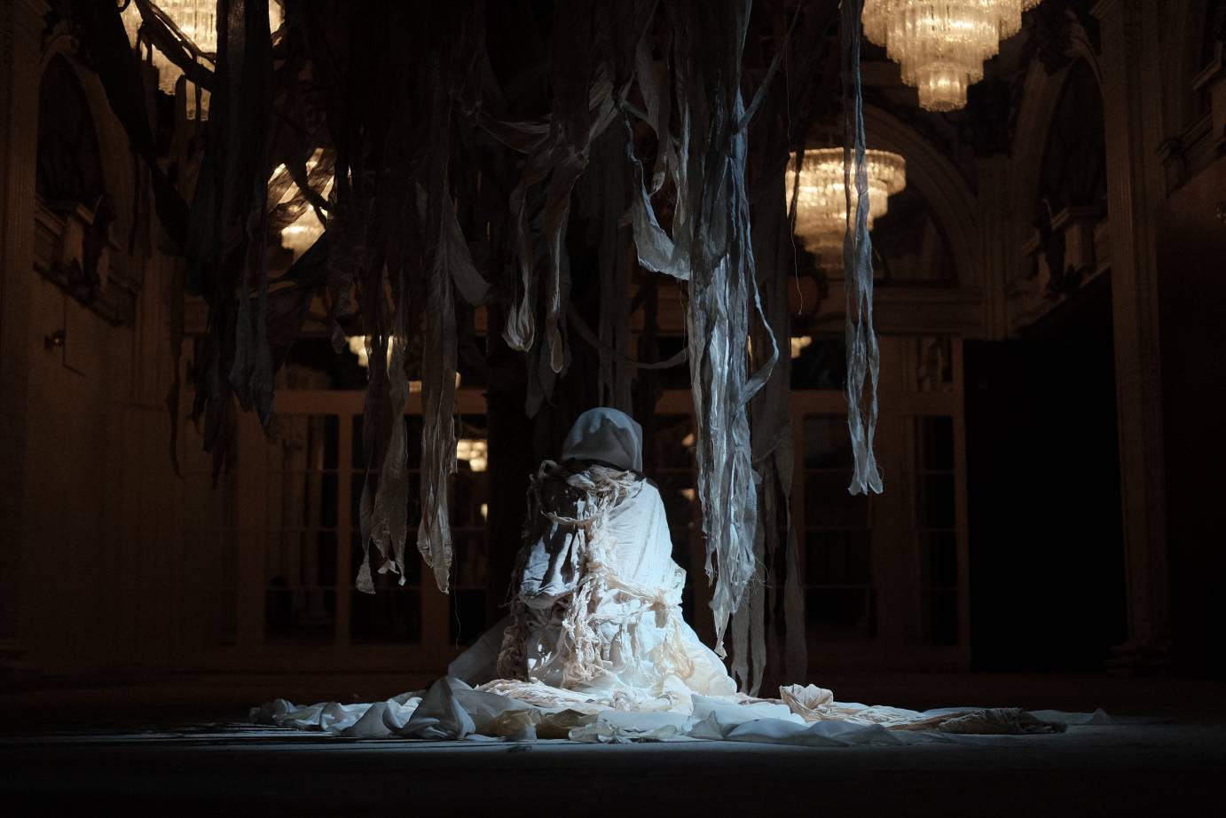 In a ballroom lit by a chandelier, fabric hangs from the ceiling as a woman dressed in rags kneels