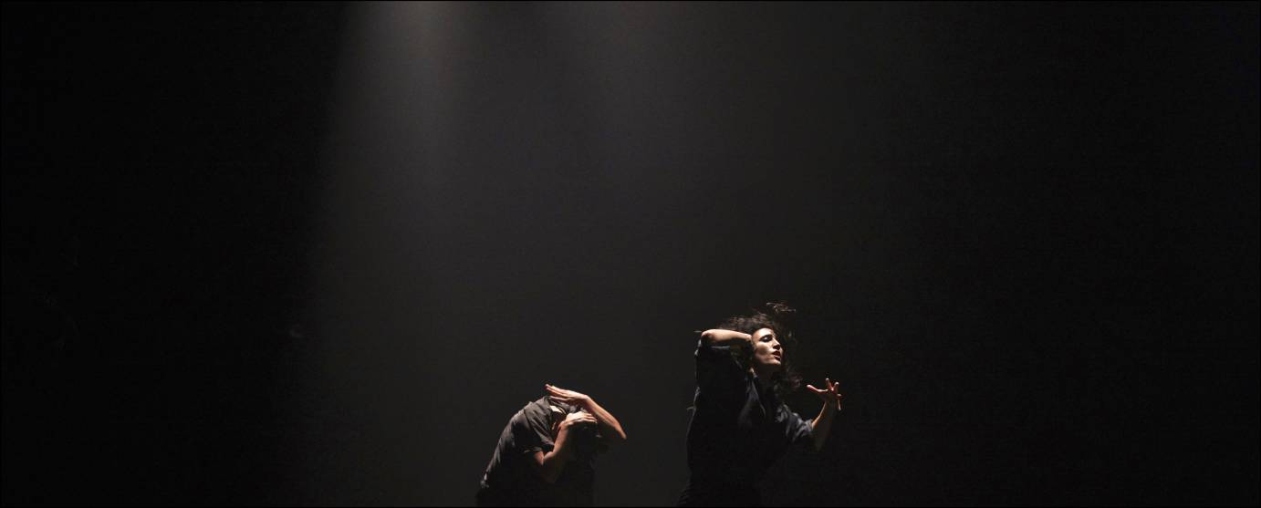 In a dark space, a man covers his head with his hands while a woman gestures