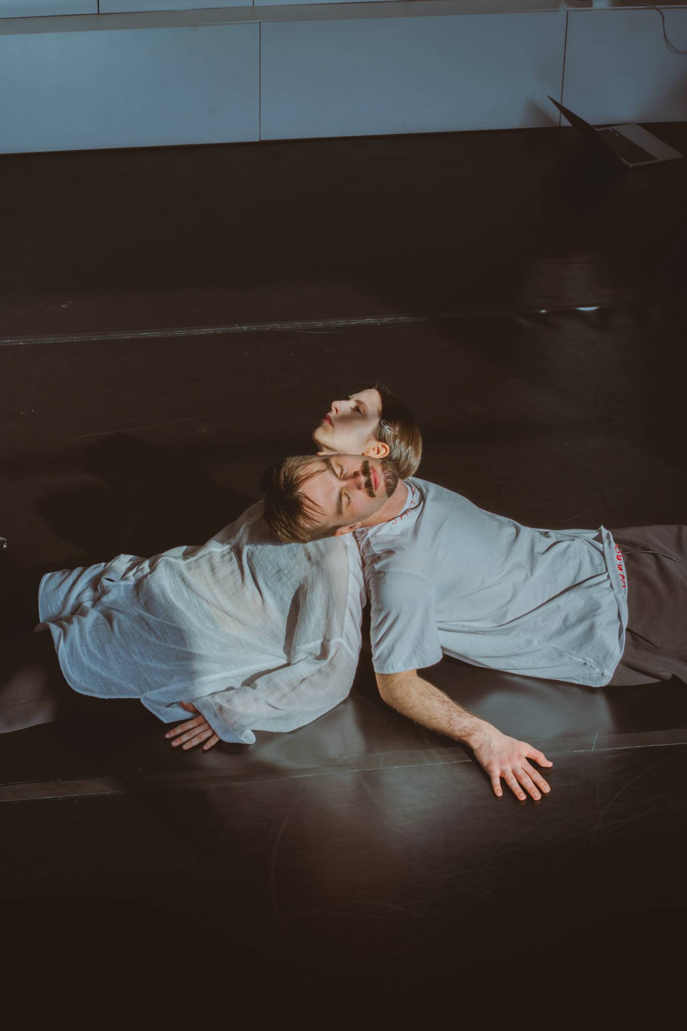 Two light-haired performers lie on the floor propped on their elbows resting their heads on the other dancer's shoulder. Both wear white T's and brown slacks. One has a mustache and beard.