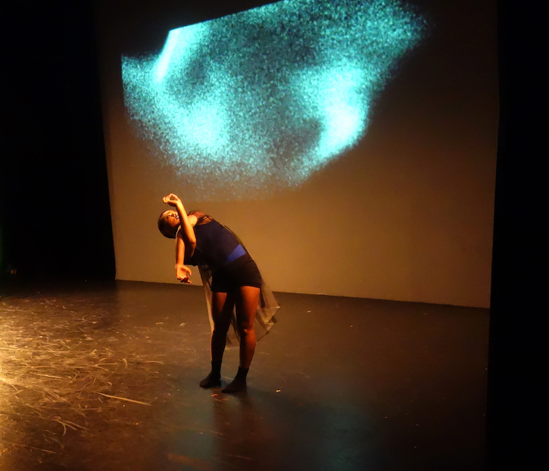 A dancer in shorts, socks and layered tops crosses her arms at her elbows and looks to the ceiling.