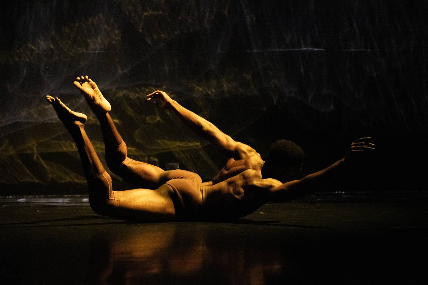 a Black man in skin matching shorts and nothing else rolls on the floor balancing on his pelvis as his legs and arms reach out and away from his torso