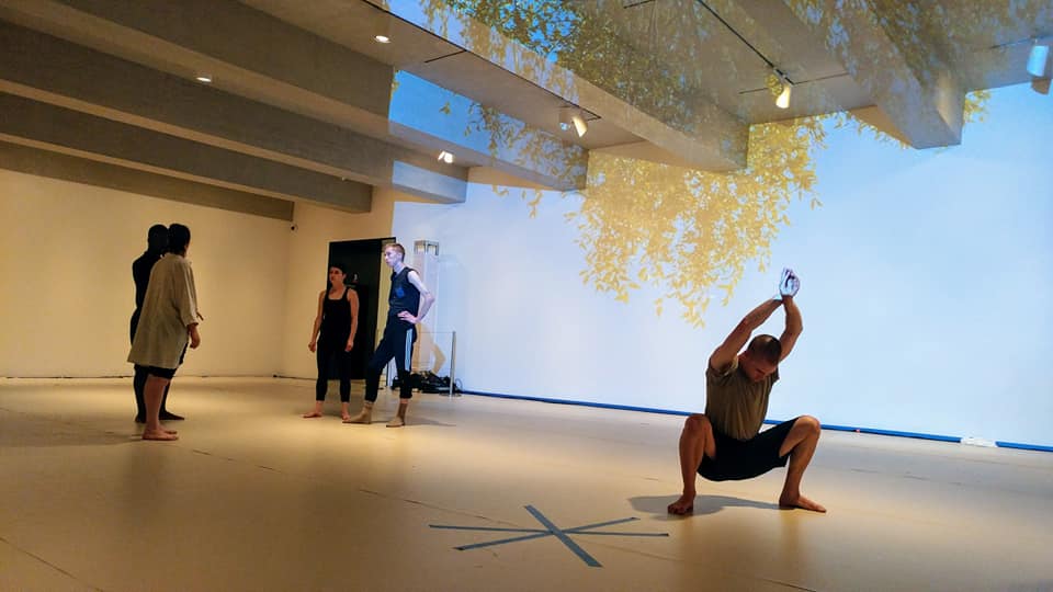 A dancer in a deep squat with his arms above his heads. A projection of a hanging branch is in the background. A group of dancers also stand in the background.