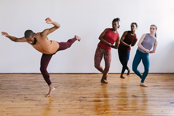 a trio of female dancers in colorful tight clothes lean back and survey a male Black man standing on one leg in an arabesque 