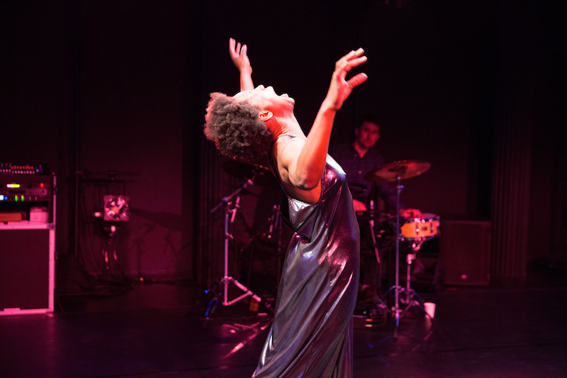 mayfield brooks wears a metallic silver gown and tosses their head back and screams out. A drummer sits near their set in the background.