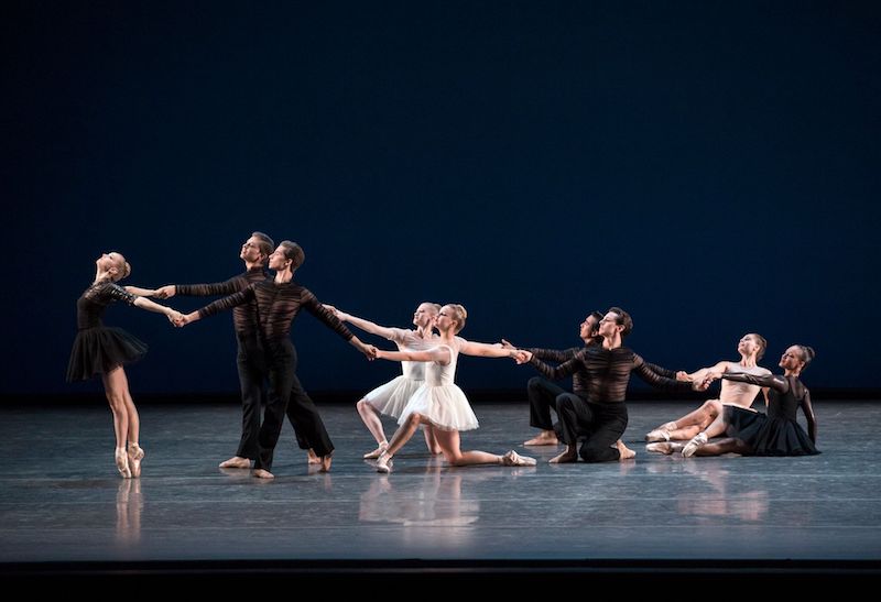 Dancers hold hands in a cascading formation in Gianna Reisen's work. Some dancers are en pointe while others sit and kneel. 