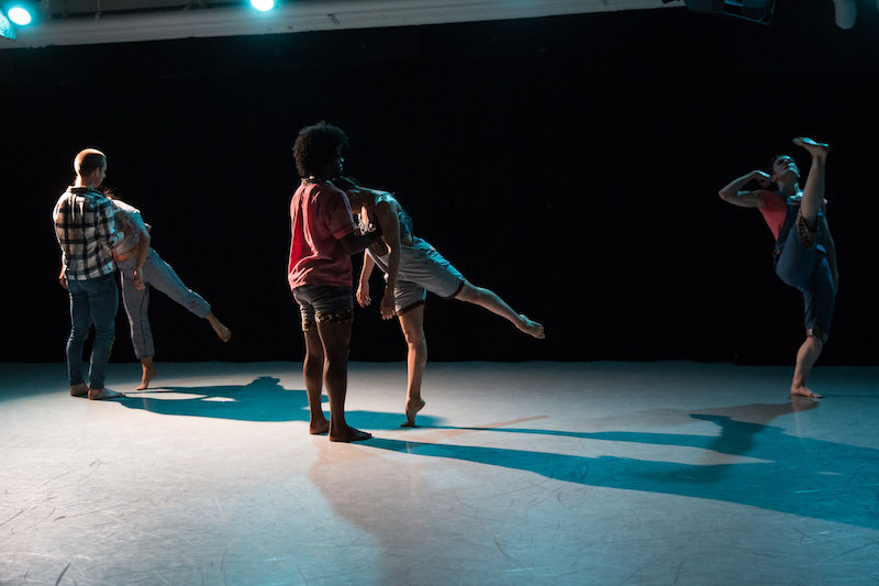 There are two couples. The women in the group arabesque and rest their heads on their male partner's chest. A woman to the right of them kicks her leg high into the sky with a flexed foot.