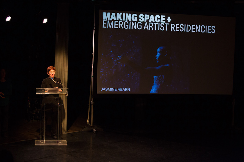 Gina Gibney stands at a podium describing the organization's new programming. A screen projections information about the new emerging art residency. A close up of artist Jasmine Hearn is also projected.