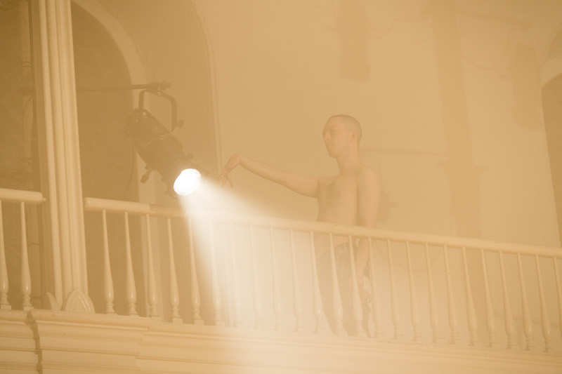 Three dancers assume positions a wood floor in midst of a hazy yellow glow. One dancer sits on a knee, another stands attention and the third assumes a warrior-like yoga post.