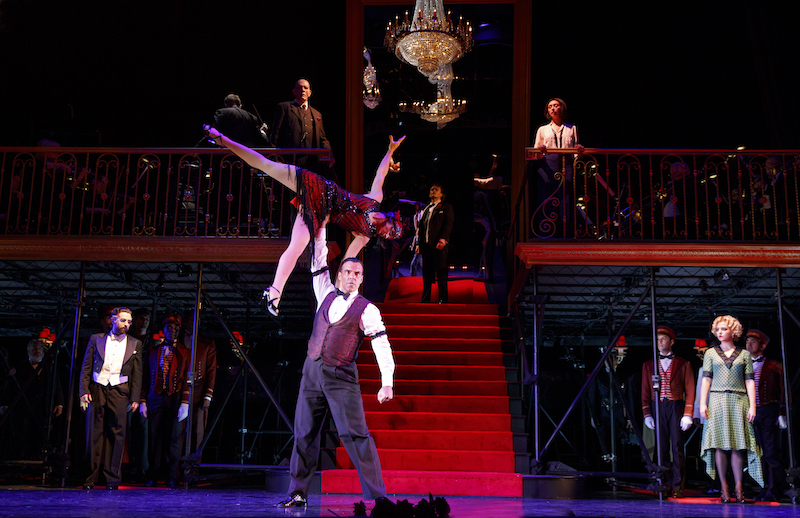 A man in shirtsleeves, red vest and slacks holds a woman above his head. The ensemble watches in the background. An ornate chandelier hangs above them.