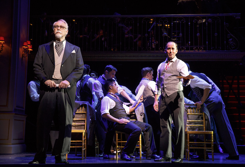 Two men in suits speaking out to the audience. A group of men in shirtsleeves sit in the background.