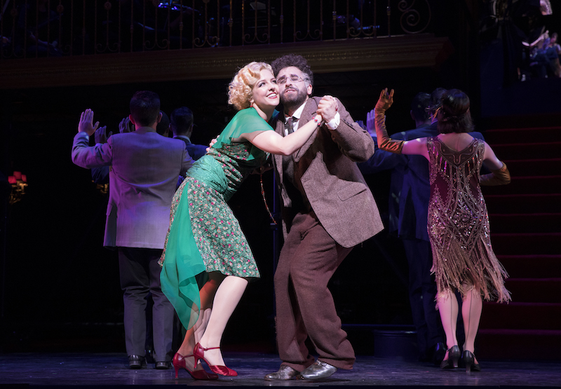 A man and woman dance cheek to cheek. The woman wears a green dress and red satin heels. The man wears a brown suit. Other couples dance in the background.