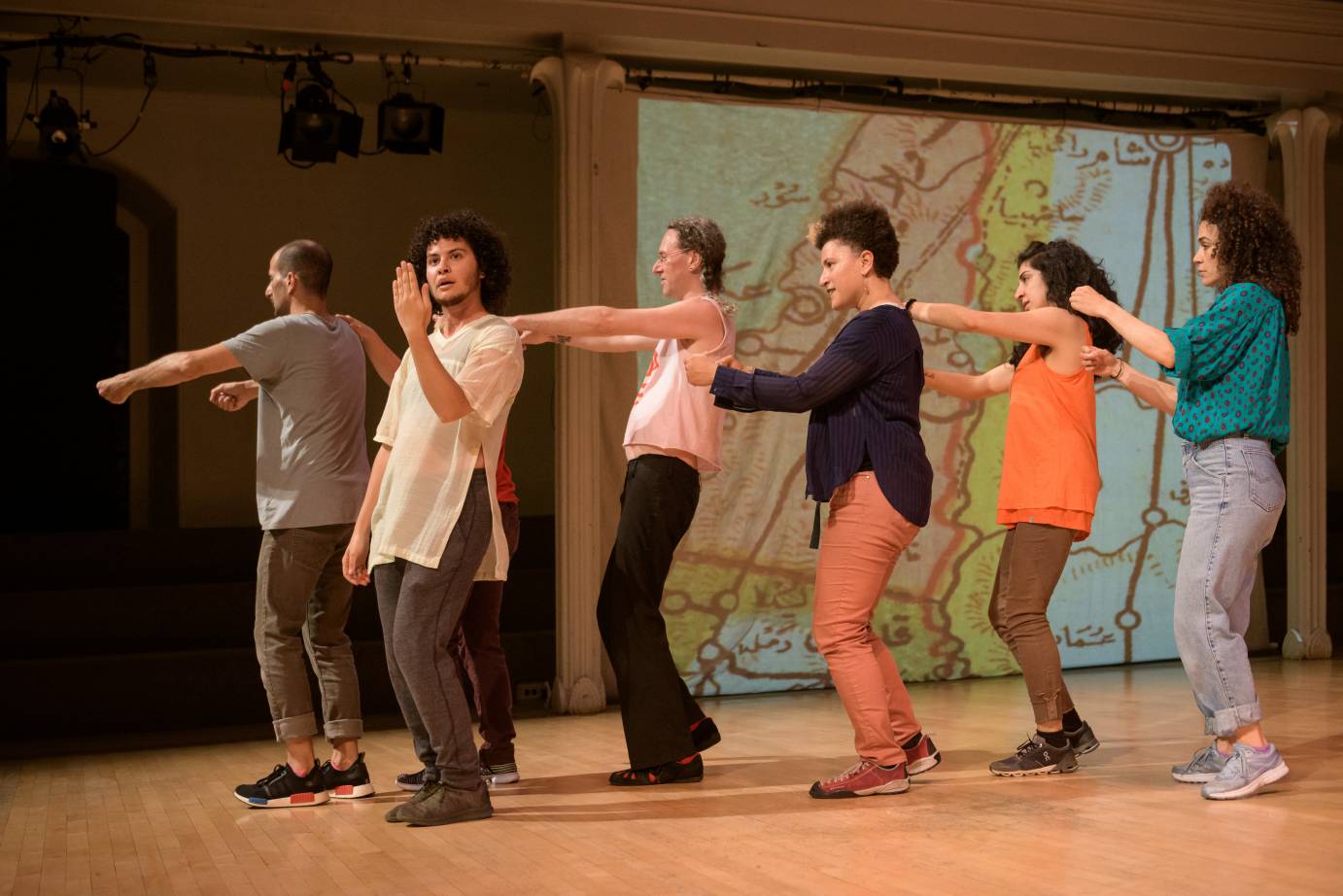 Dancers hold each other's shoulders in a line while one man lifts his hand