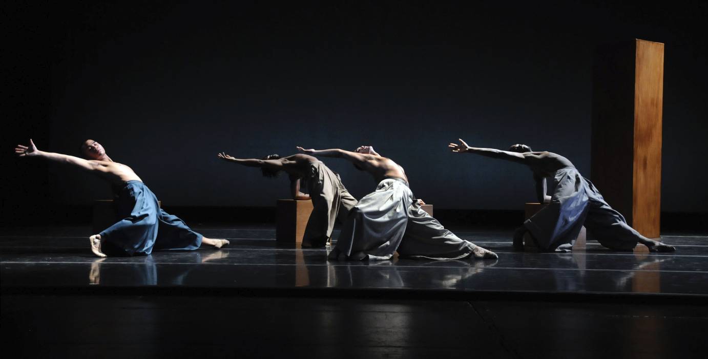 Shirtless male dancers are in a grounded stag pose, one arms extendedarm