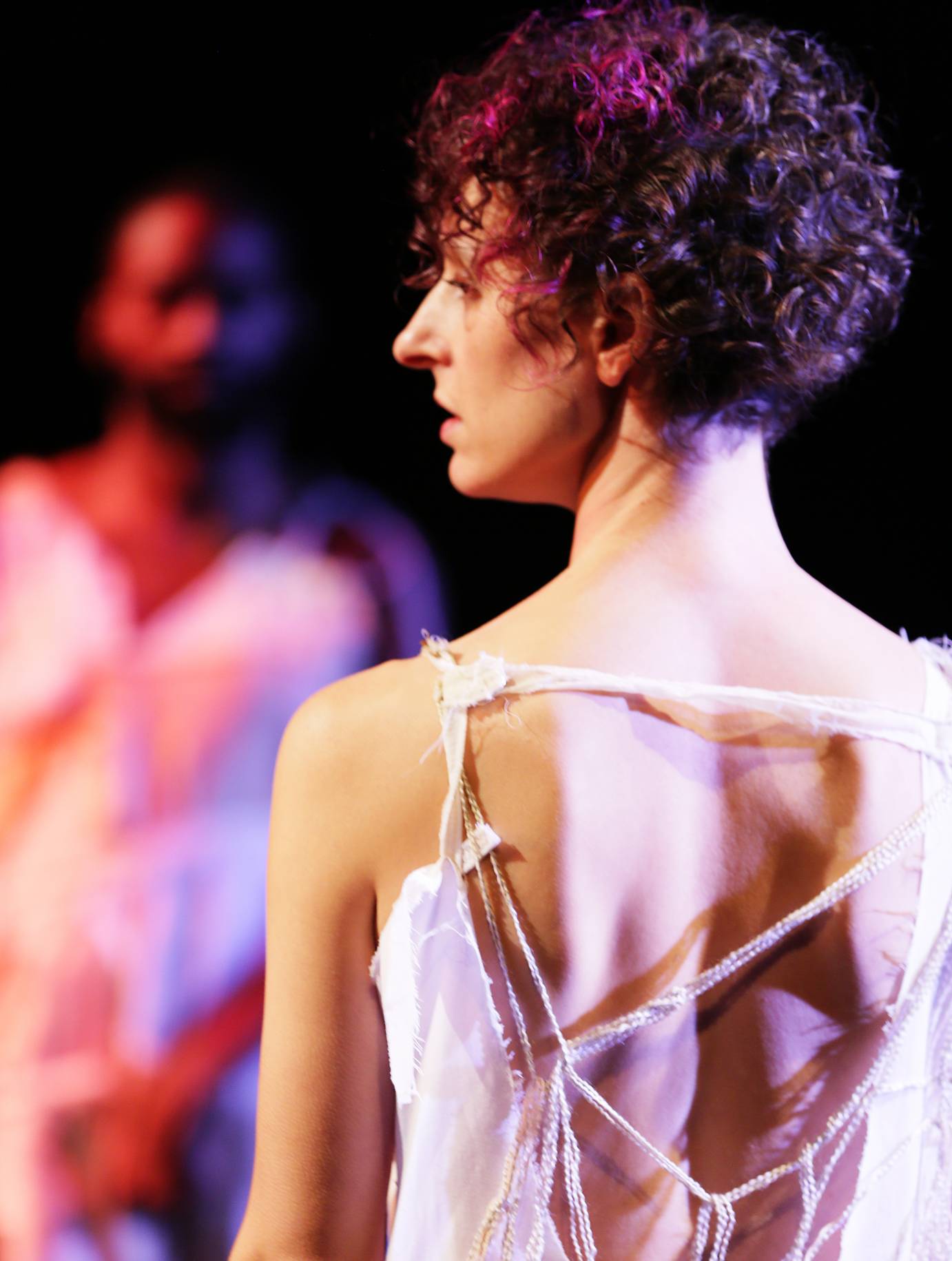 A profile of a female dancer with curly hair; another man is blurred in the background