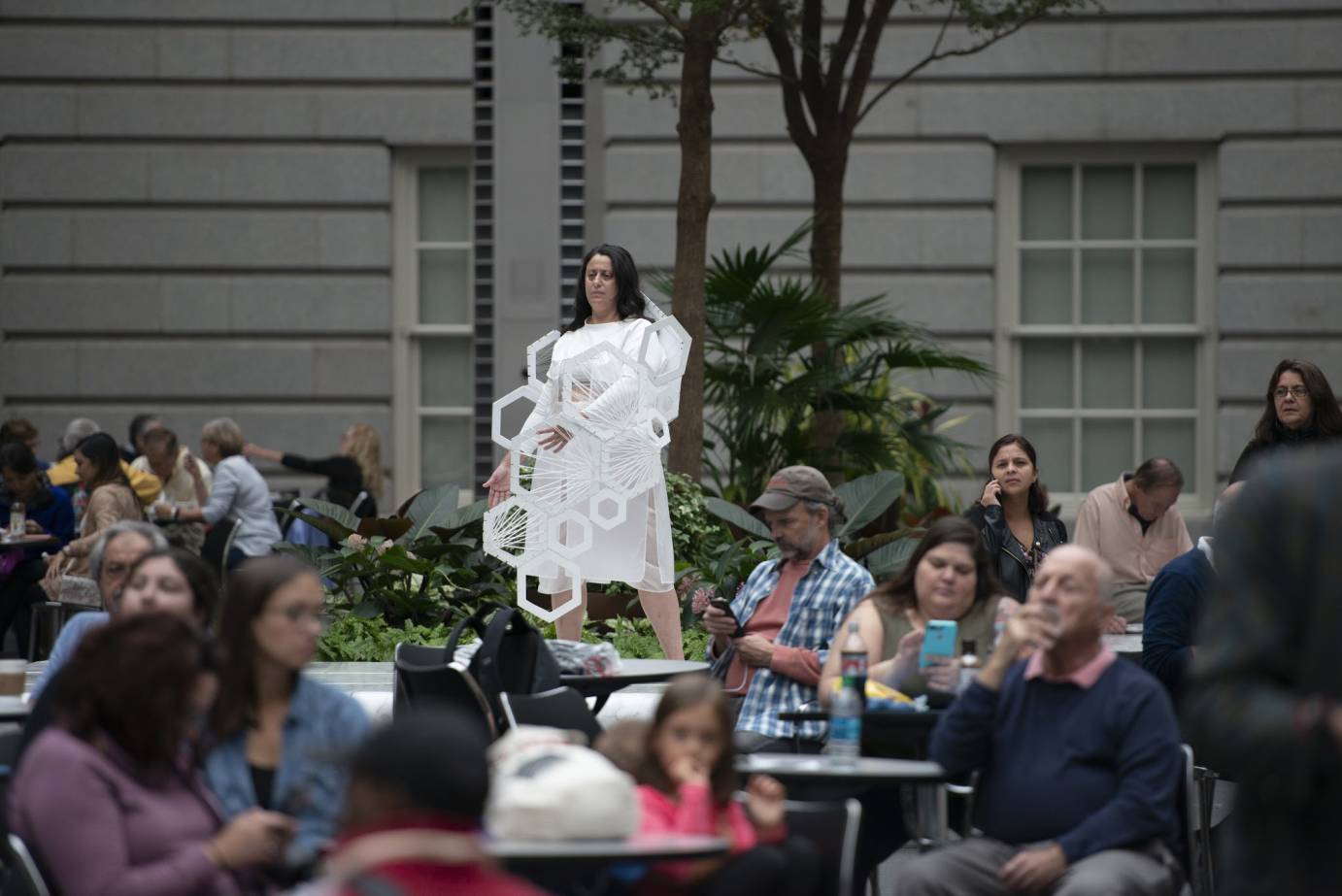 A woman in white stands on a platform as pedestrians around her look at their phones and eat lunch