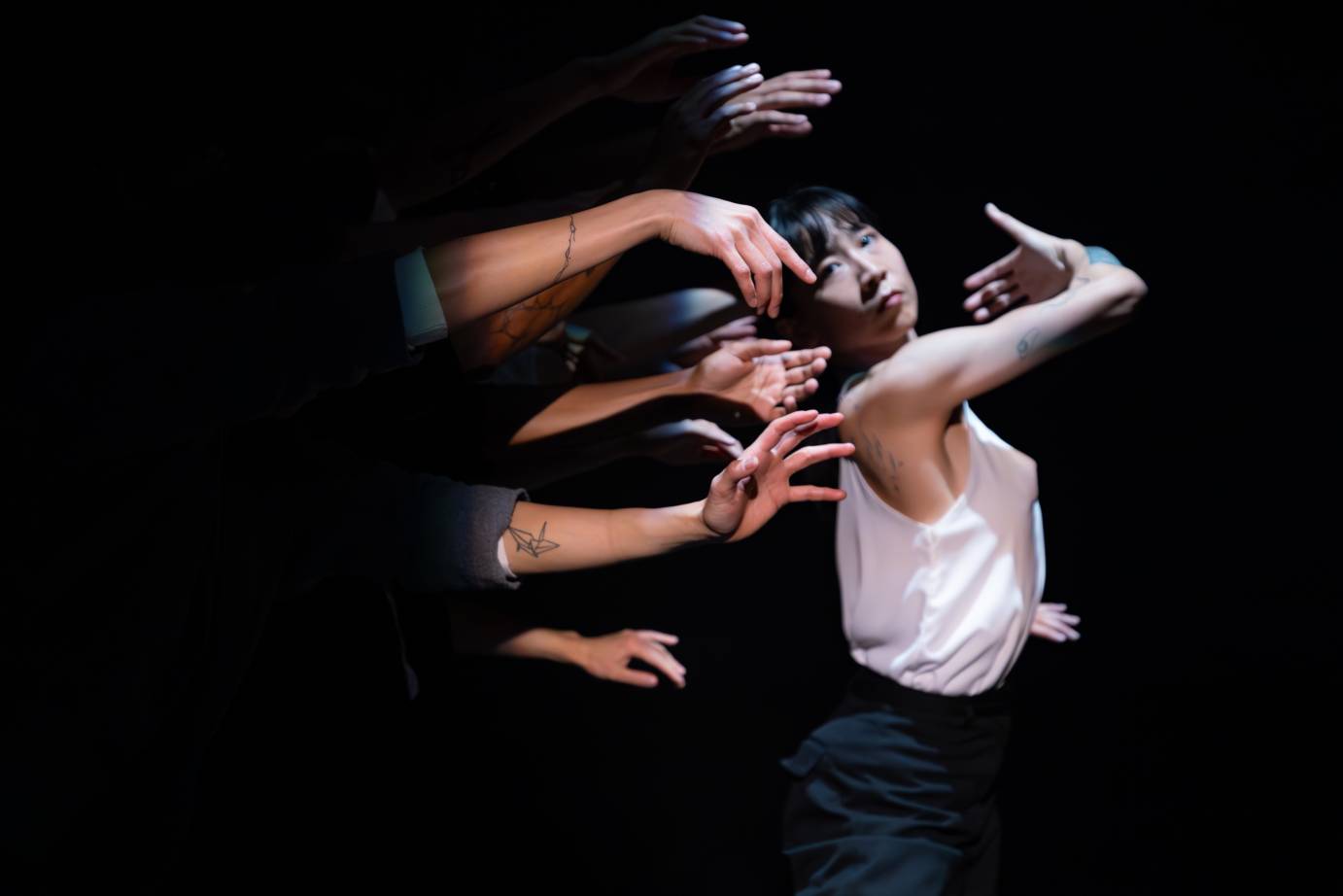 A woman seems to be leaning her back as she stands upright into the disembodied hands of several dancers.