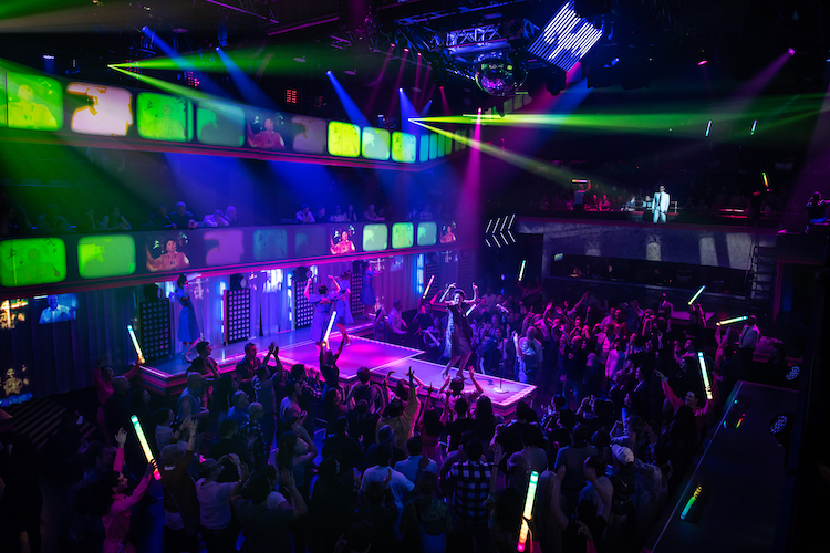 The Imelda Marcos character dances in the theatre turned disco, green and purple screens with images of Imelda Marcos fill a wall behind her.. the audience stands on the floor and dances... some holding green glowsticks.