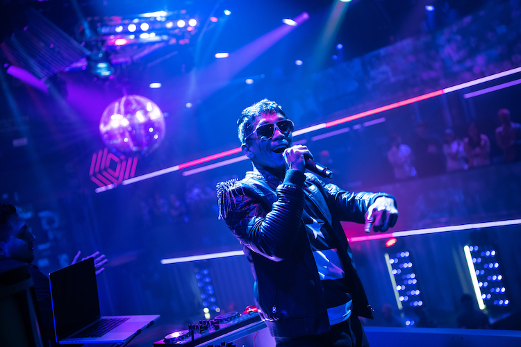 the dj is pictured hear, the lighting around him is magenta, he speaks or signs into a mic and wears aviator sun glasses and a fabulous leather jacket. behind him is a big disco ball. He is having fun.