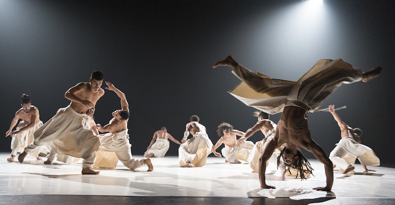 A group of bare-chested men in white flow-y pants spiral into the ground. One in the foreground is upside down in mid cartwheel. 