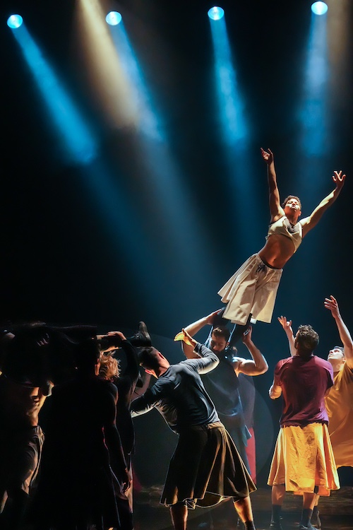the company surround a dancer who stands on top of her castmates shoulders as she leans precariously to one side.