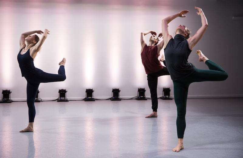 Three dancers stand on leg. Their other legs are bent in back attitude positions while their arms are thrust behind them and their backs arch back. They look up to the heavens.