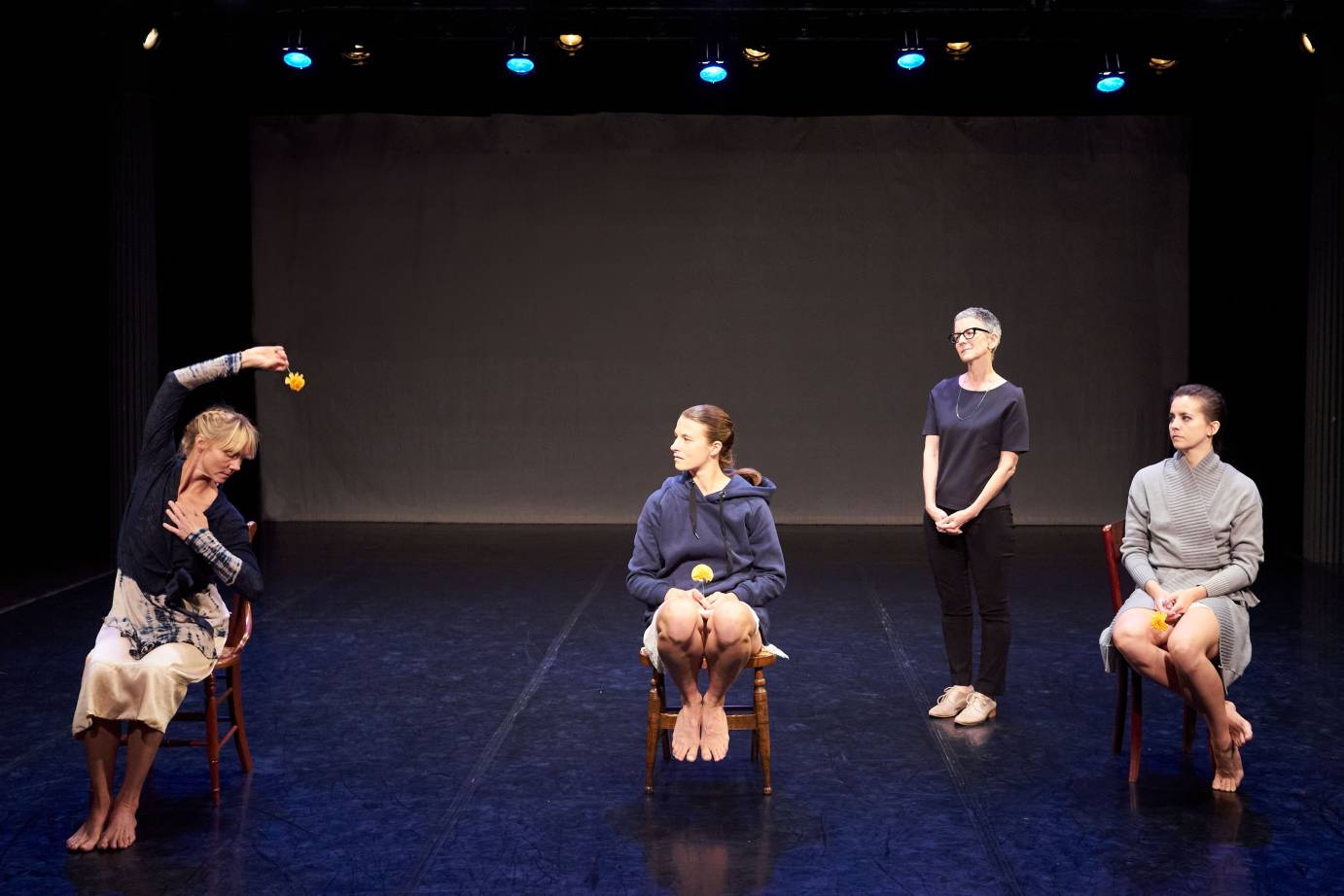 Four women, three seated and one standing, watch the dancer on their left frame her head with a marigold flower suspended by her right hand.