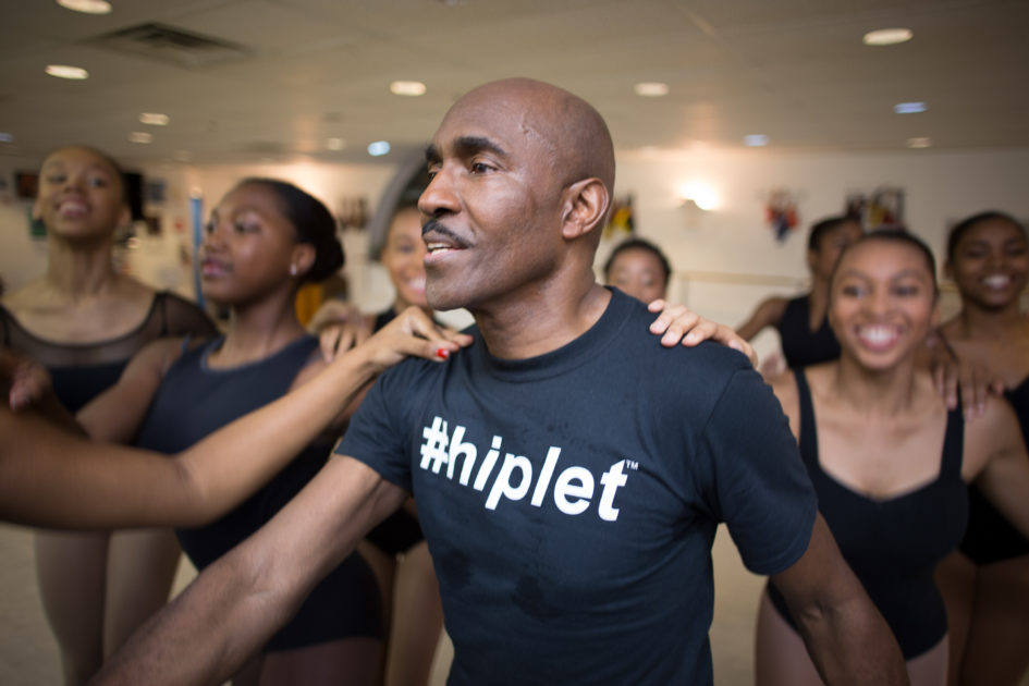 Homer Bryant a black man in a dance studio with his black tshirt, inscribed with hiplet, and his laughing  students in the background