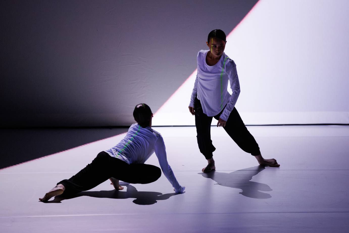 Two women in close proximity, one in a crawling position looking up at the other dancer, while the other, standing dancer, spirals and looks away from the dancer below.