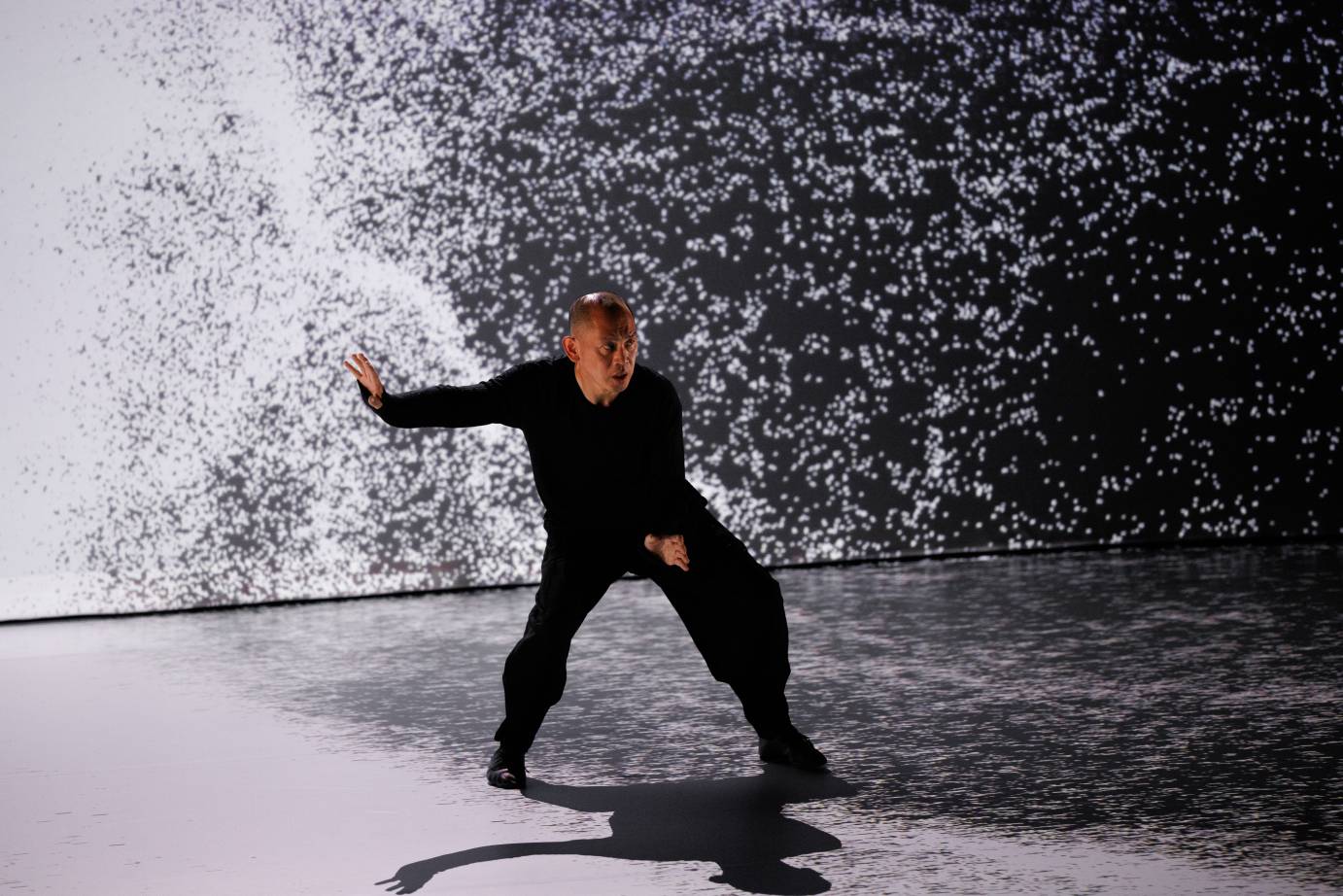 A man with an intense facial expression looks out as he seems to be bombarded with black and white projections on the stage floor and behind him. 