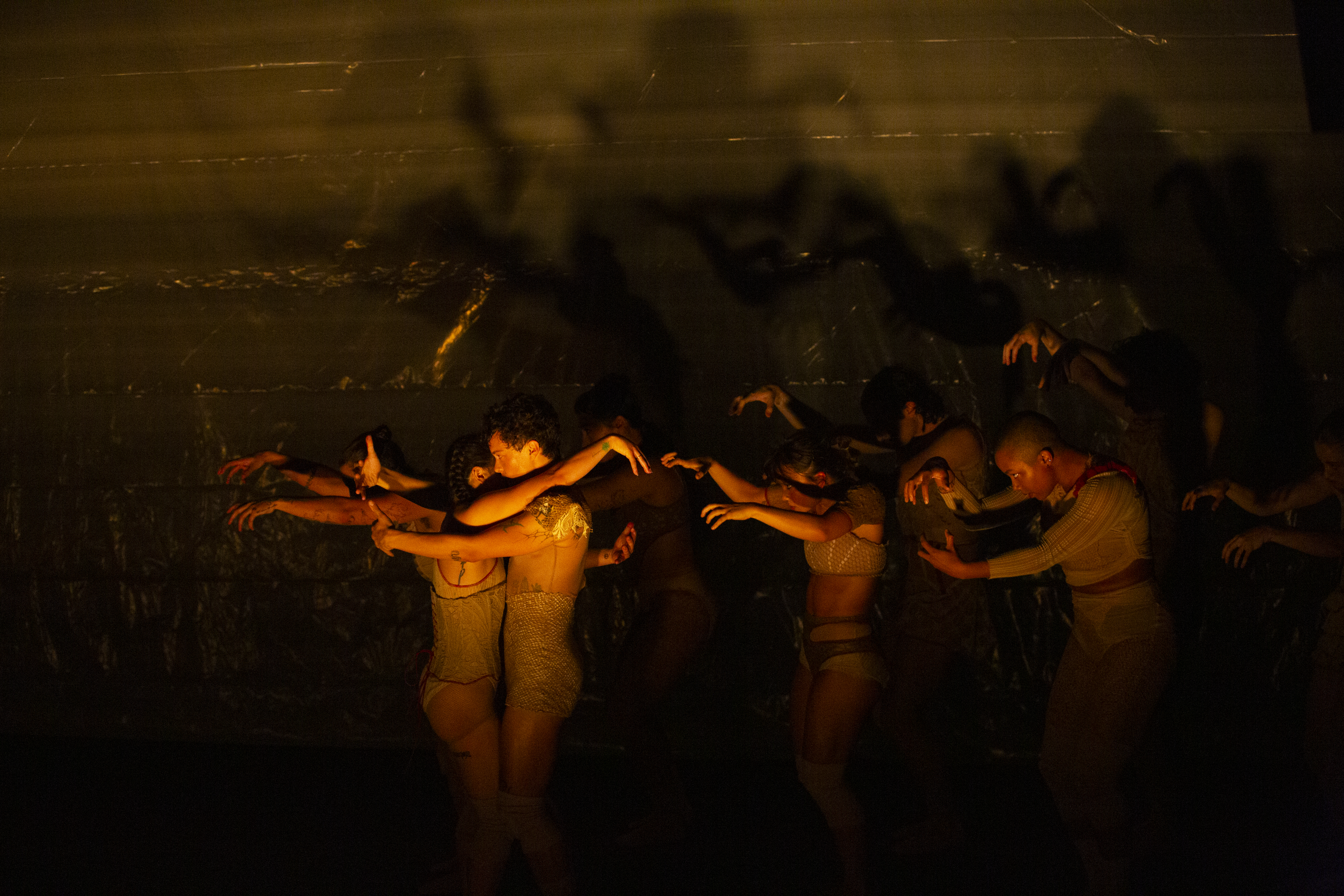 The upper bodies of several dancers are illuminated in a warm glow while the remainder of the stage is dark. One couple loosely wraps their arms around one another while others mimic the wrapping but there is no partner.