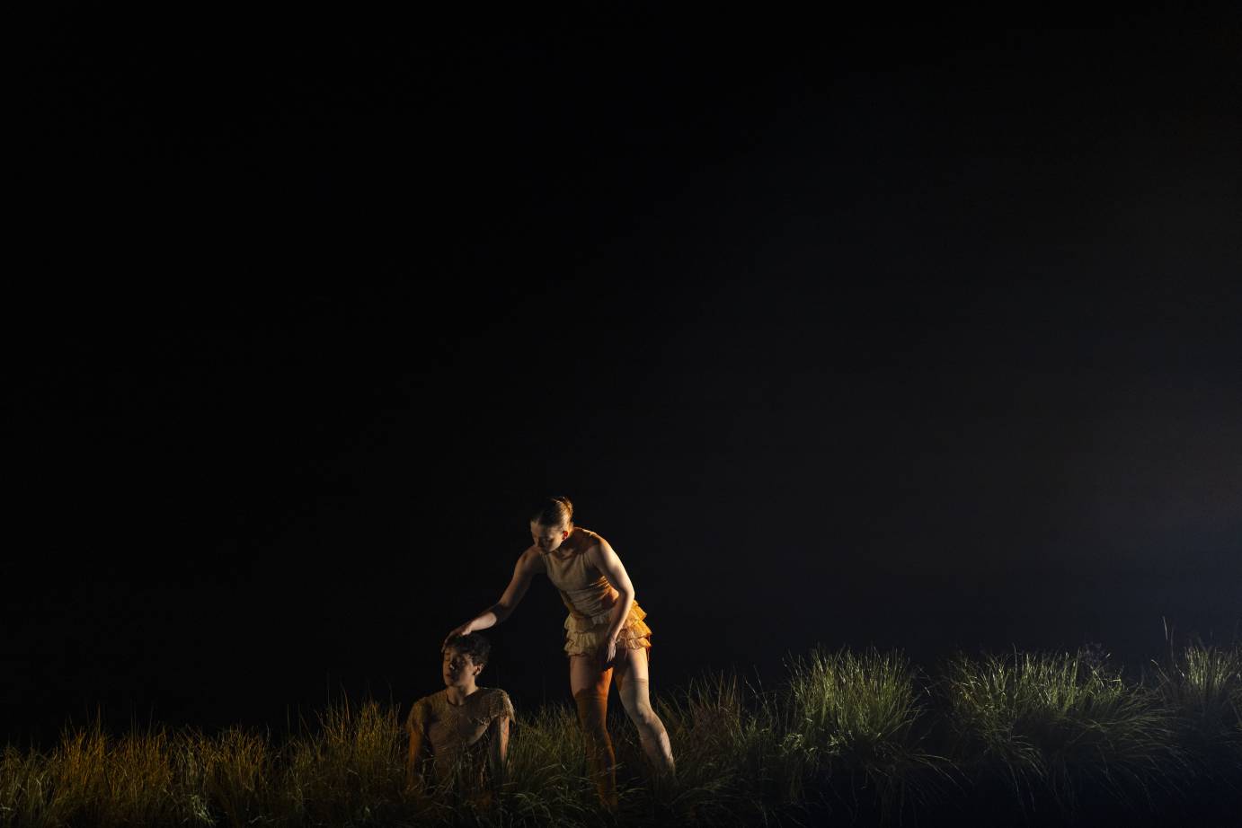 A woman stands as she places her hand on the head of a sitting man. She looks down at him while hie looks out. Thigh high blades of grass form the backdrop.