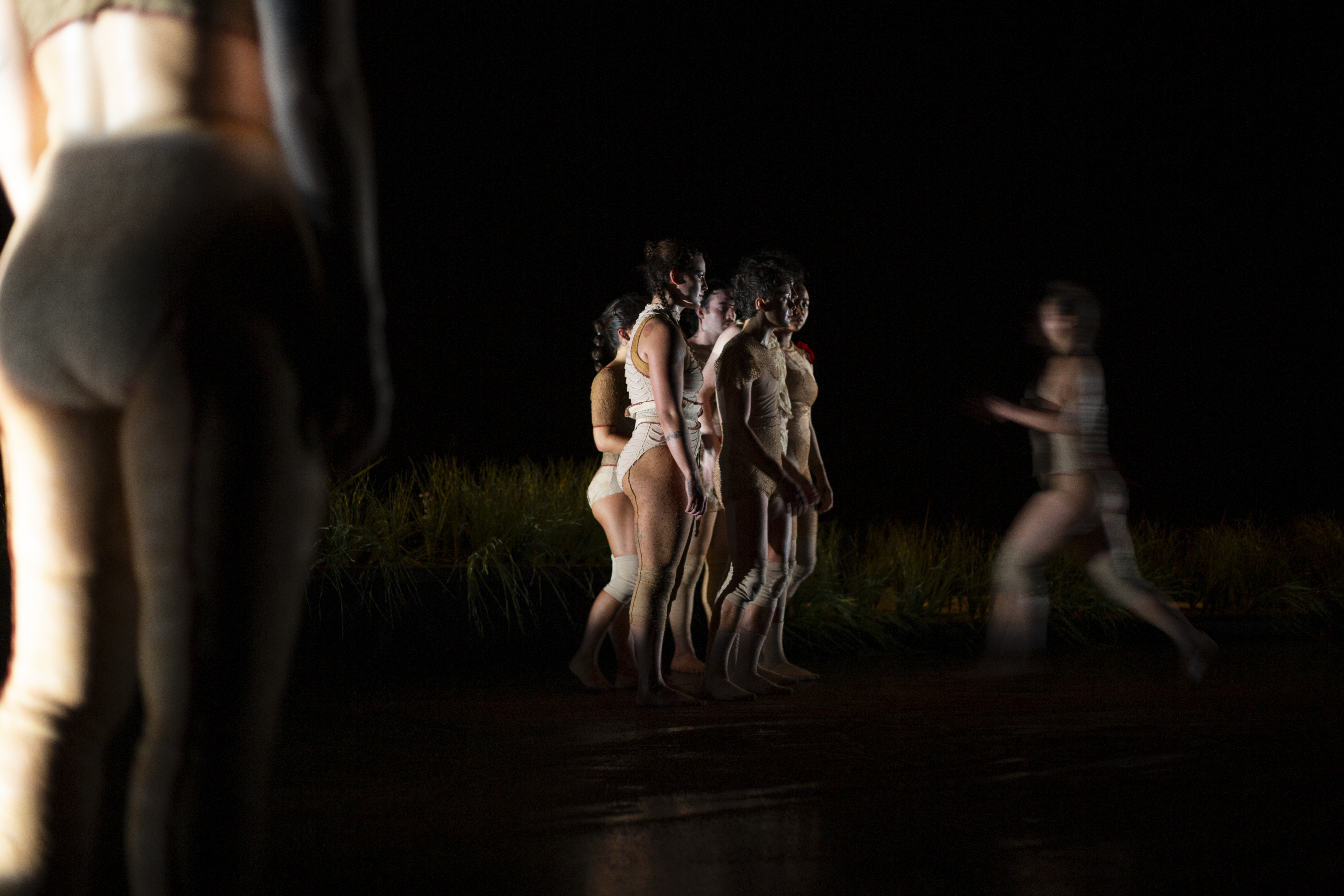 A group of women dancers in white leotards with red and blue detailing clump together. A dancer's backend and legs are enlarged on the left side of the photo.