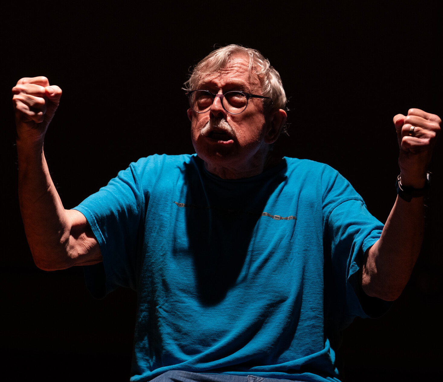 A bespeckled white haired mustachioed man dressed in a bright blue T-shirt raises his fists.