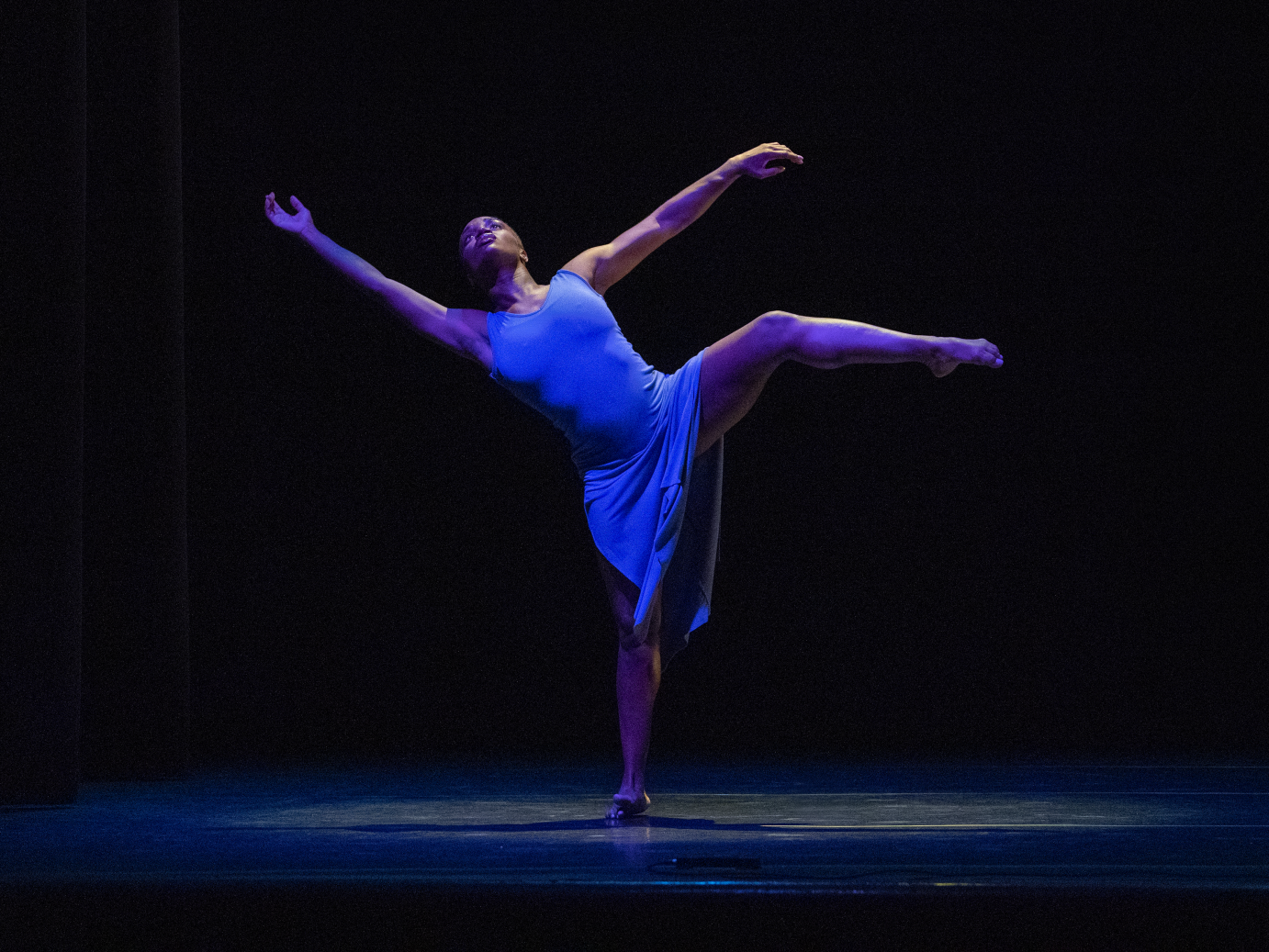 A woman in a blue tank dress lifts her leg in a side attitude. She leans away from the leg, her arms extended upward.