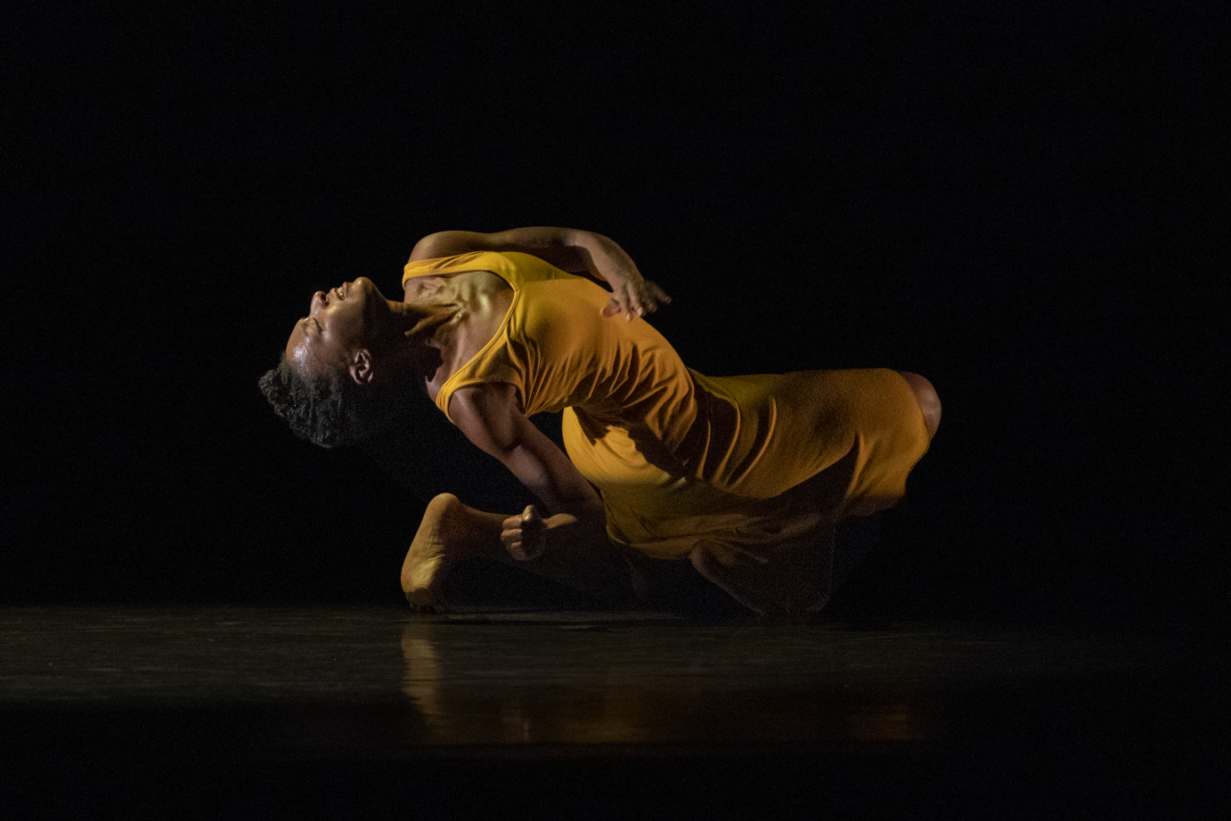 A woman in a yellow tank dress crouches on the floor, leaning backward until she is looking skyward.