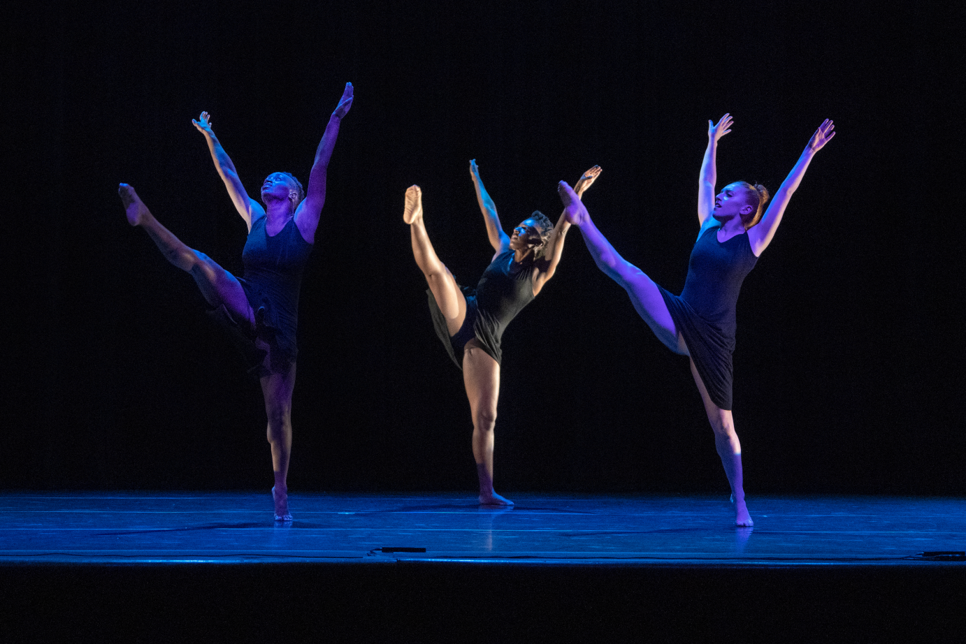 Three women extended their arms upwards while executing a high kick forward. They are facing the front corner of the performing space.