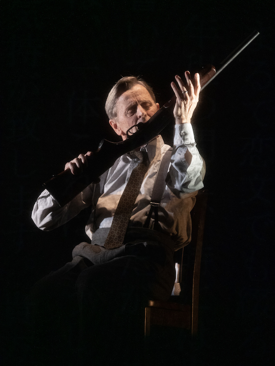 M. Baryshnikov in a crisp oxford shirt with brown pants held up by suspenders sits and intently cleanse a rifle. 