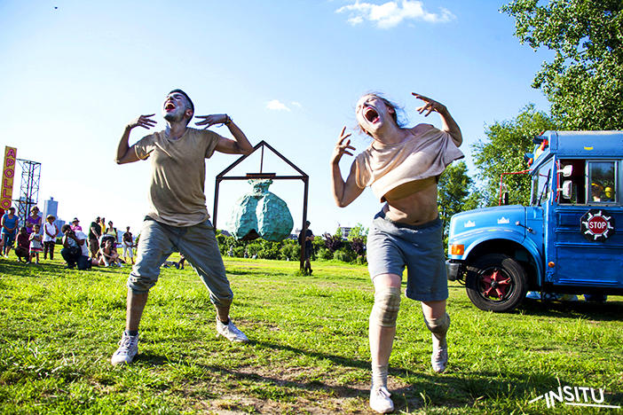 Two dancers wearing white face paint make faces. The backdrop sees an attentive audience and a blue school bus.