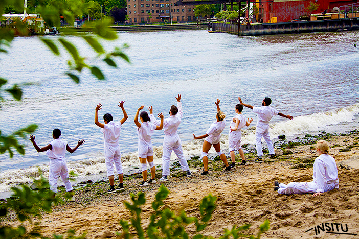 Seven performers dressed in all-white stand on the edge of a beach with their arms outstretched. A solo observer is seated on the sand behind them.