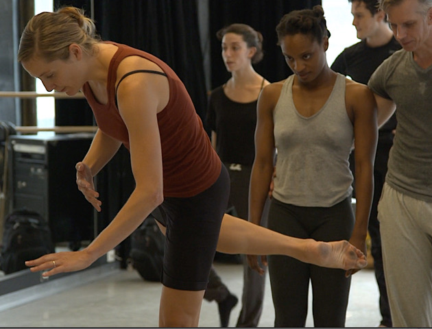 A woman stands in an attitude position as other dancers observe her directions