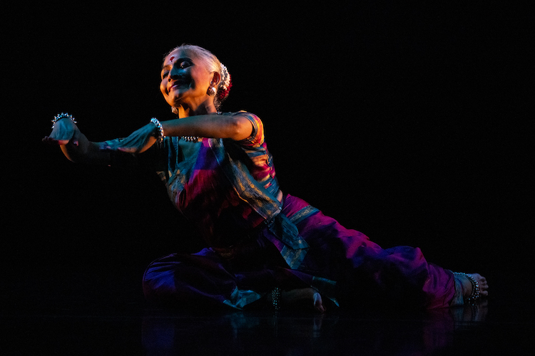 Bijayini Satpathy sits on the floor one leg bent in front of her , the other behind her. The focus is on her illuminated face and her hands. She seems to be telling a story with her hands and appears to be enjoying it.